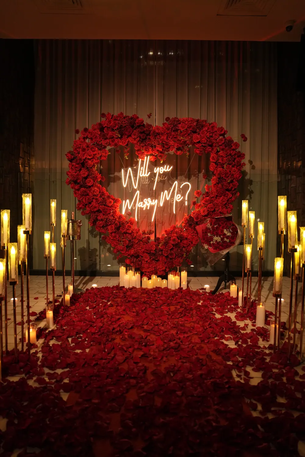 A red rose heart with a glowing Will You Marry Me sign, framed by tall gold candle stands over a floor of red petals