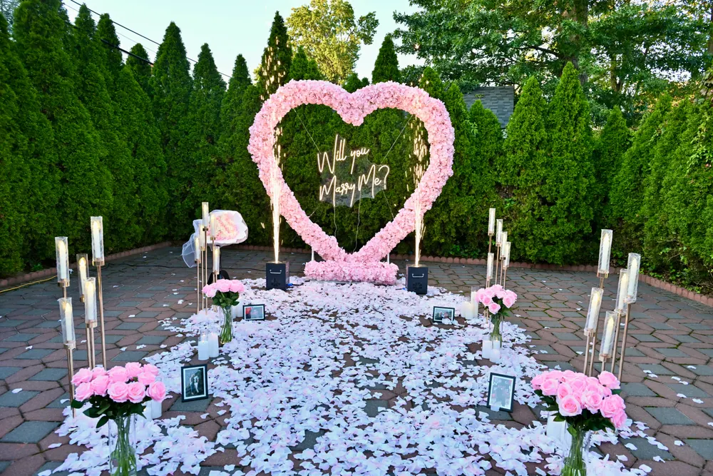 A pink rose heart with a Will You Marry Me sign in a garden courtyard, with white and lilac petals, pink roses and framed photographs