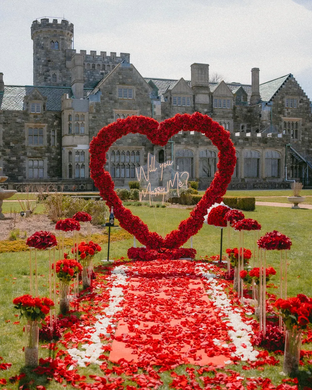 A red rose heart and petal aisle on the lawn of a stone estate, lined with red roses and candles