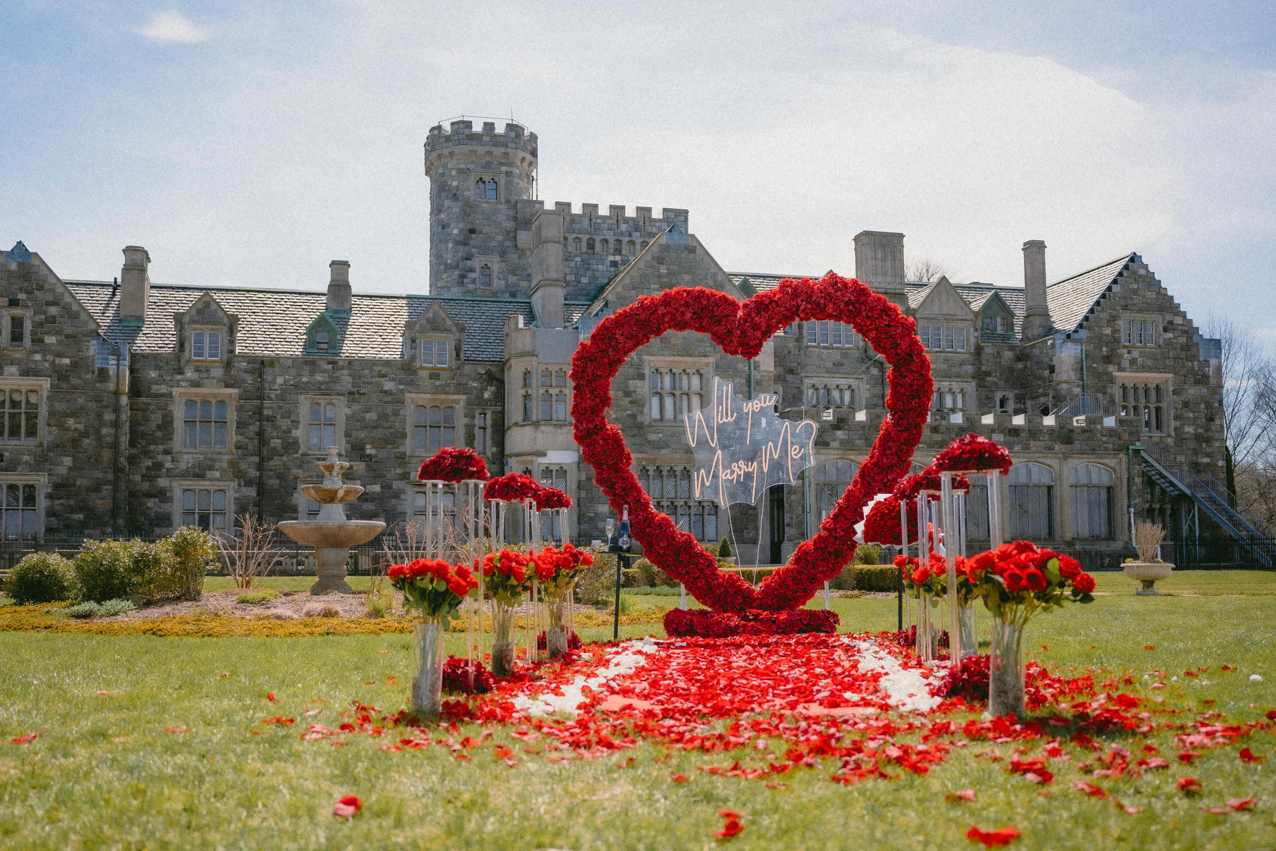 A red rose heart with a neon Will You Marry Me sign on the lawn of a stone castle estate, red roses on clear stands lining a petal aisle