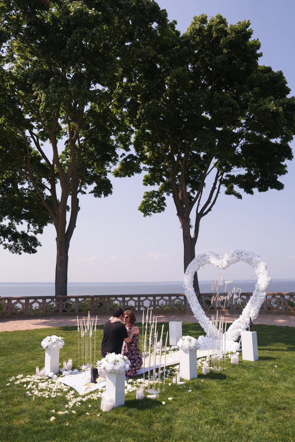 A white floral heart on a lawn under two tall trees by the water, candles along a white petal aisle
