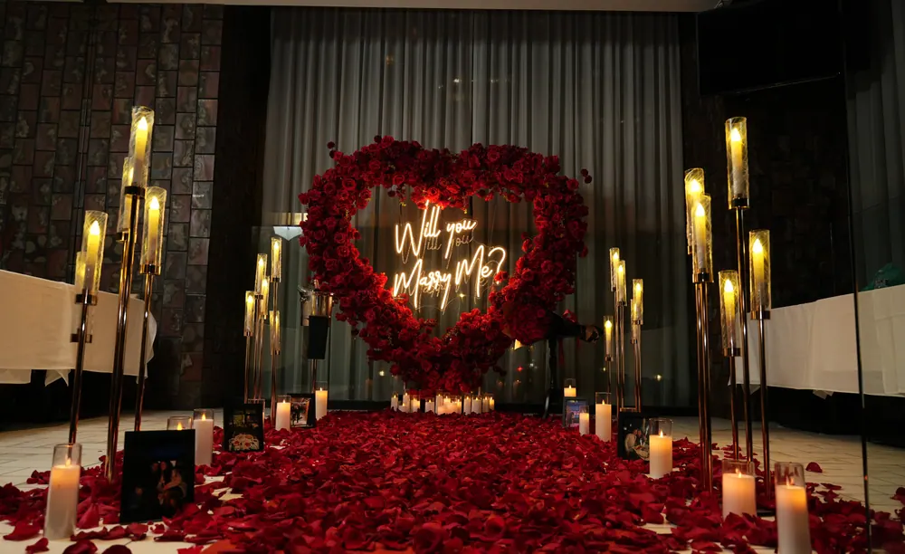 A red rose heart with a warm neon Will You Marry Me sign in a dark room, lit by tall candle stands and a floor of red petals