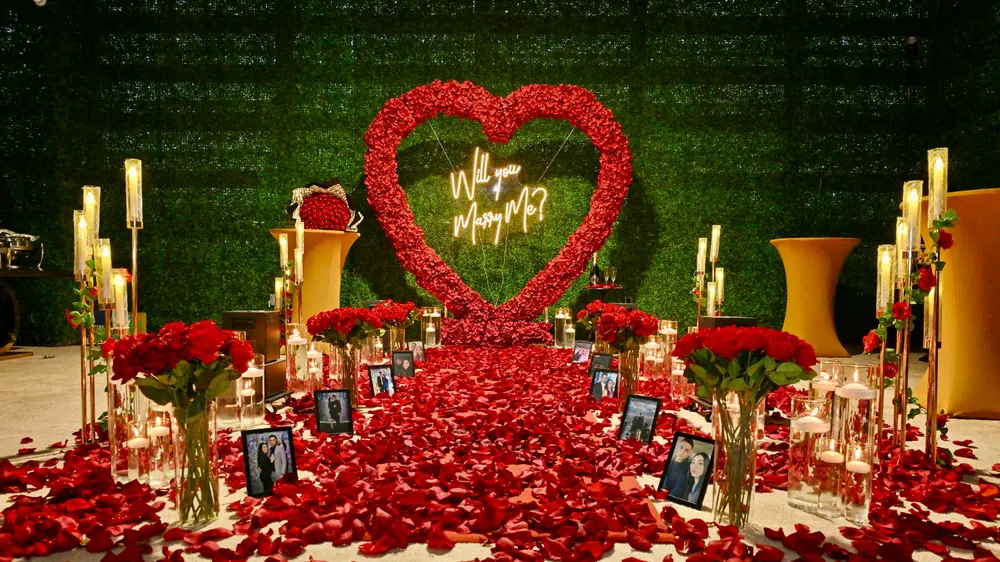 An intimate setup: a red rose heart on a greenery wall with candles, vases of roses and framed photographs of the couple