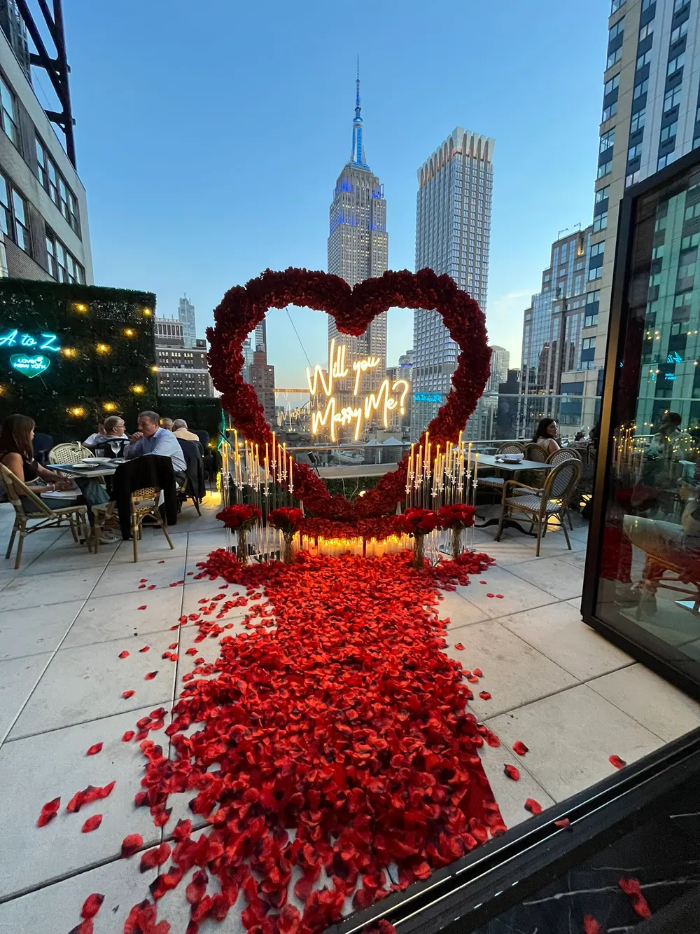A red rose heart with a lit Will You Marry Me sign on a Manhattan rooftop terrace, a rose-petal aisle leading to it and the Empire State Building behind
