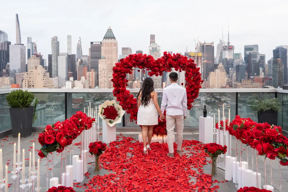 A couple standing inside a red rose heart on a Manhattan rooftop, surrounded by roses, petals and candles
