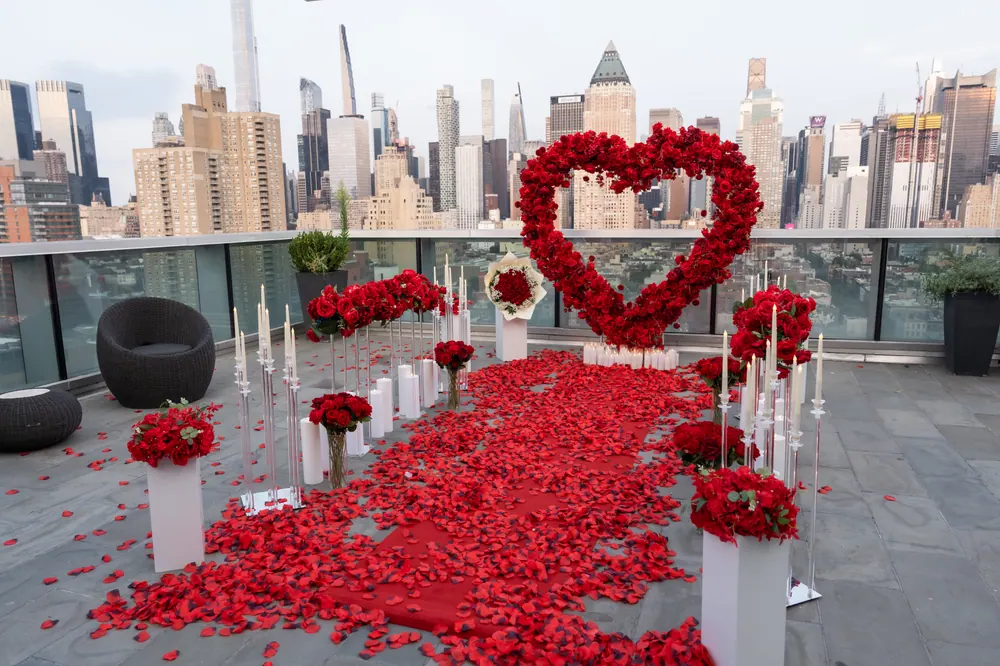 A red rose heart and petal aisle on a rooftop overlooking the Manhattan skyline, with white pillars of roses and candles