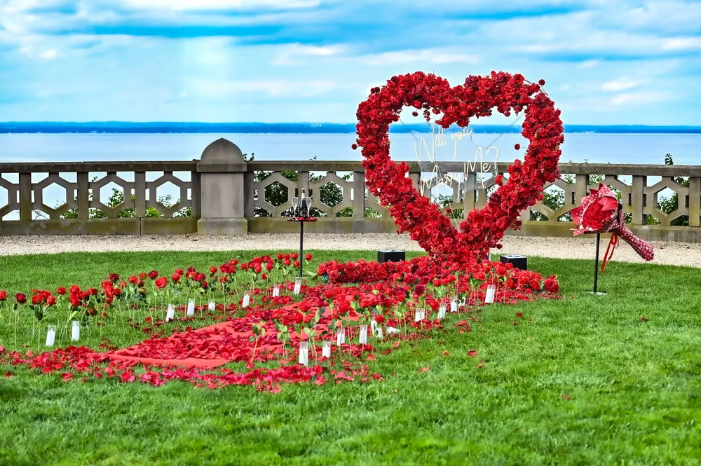 A red rose heart on a waterfront lawn beside a stone balustrade, with a winding path of red petals and candles