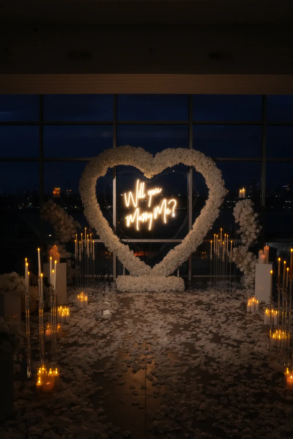 A white rose heart with a neon Will You Marry Me sign in front of floor-to-ceiling windows at night, surrounded by candles and white petals