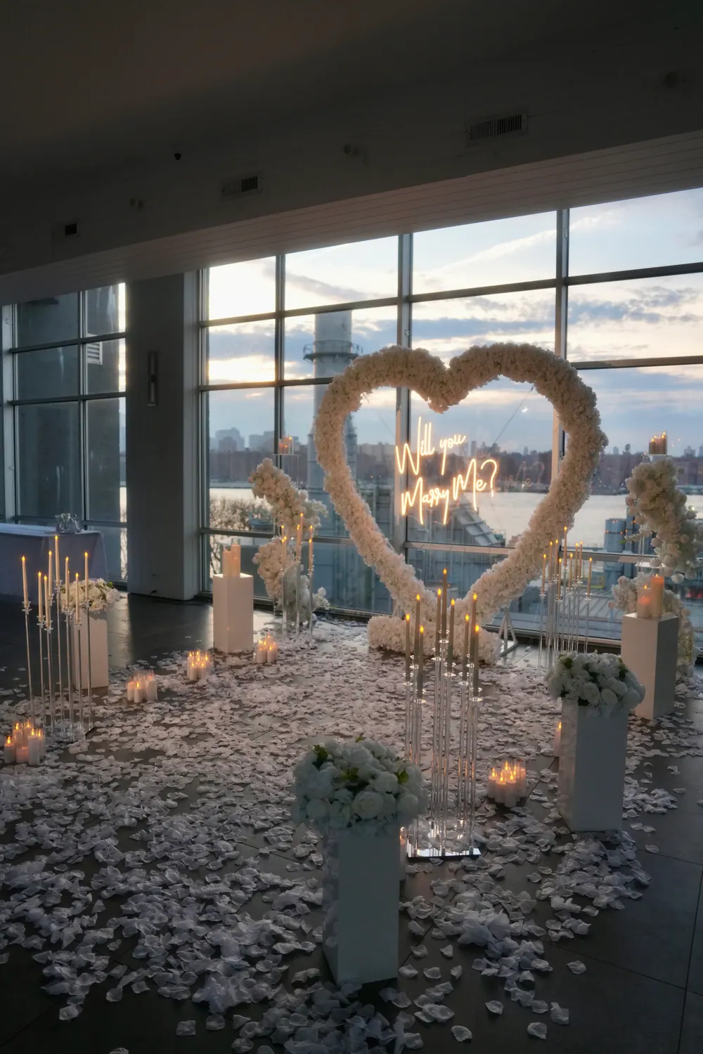 A white rose heart with a Will You Marry Me sign by tall windows overlooking the river, with white petals, candles and floral pillars