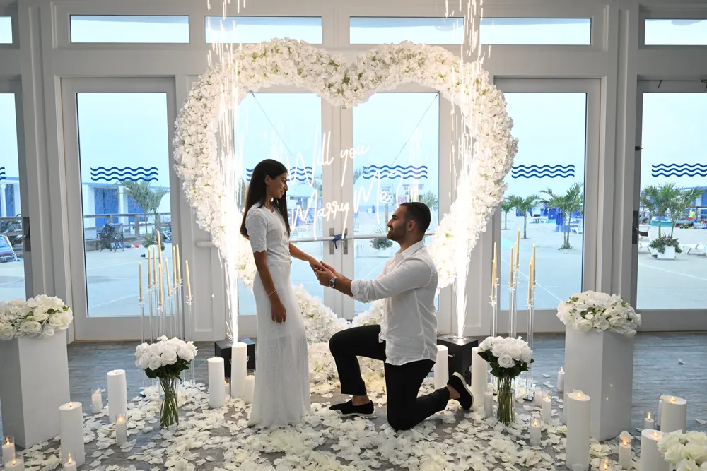 A man on one knee in front of a white floral heart in a bright glass-walled venue