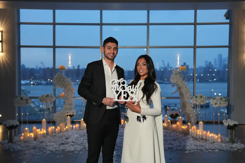 A couple holding a She Said Yes sign in a glass-walled venue at dusk
