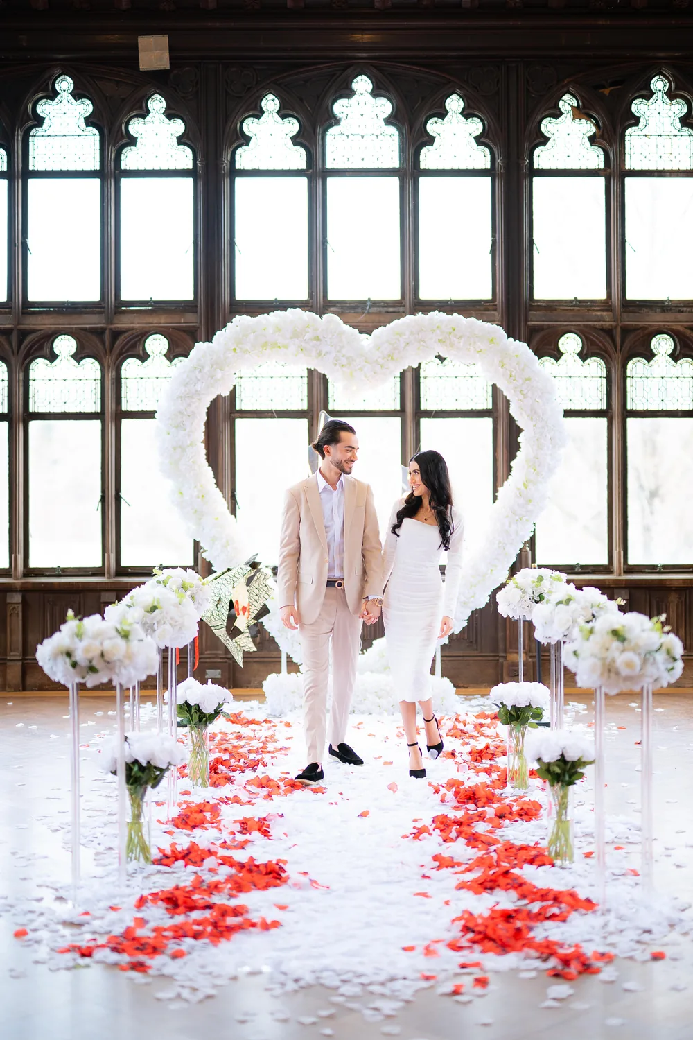 A couple standing inside a white floral heart in a panelled hall, a red and white petal aisle in front of them