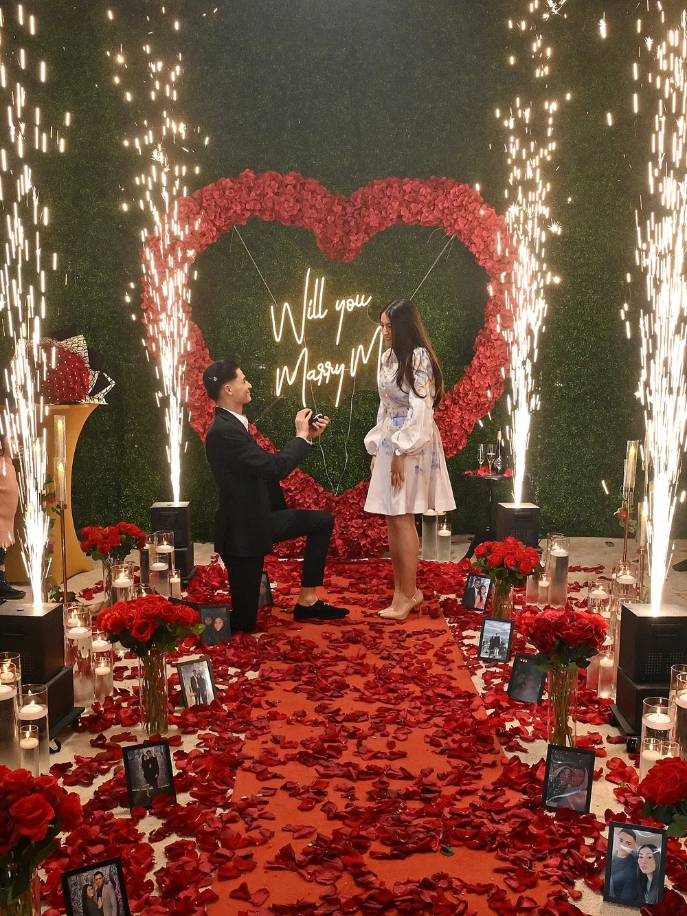 A couple in front of a red rose heart on a hedge wall, sparkler fountains either side