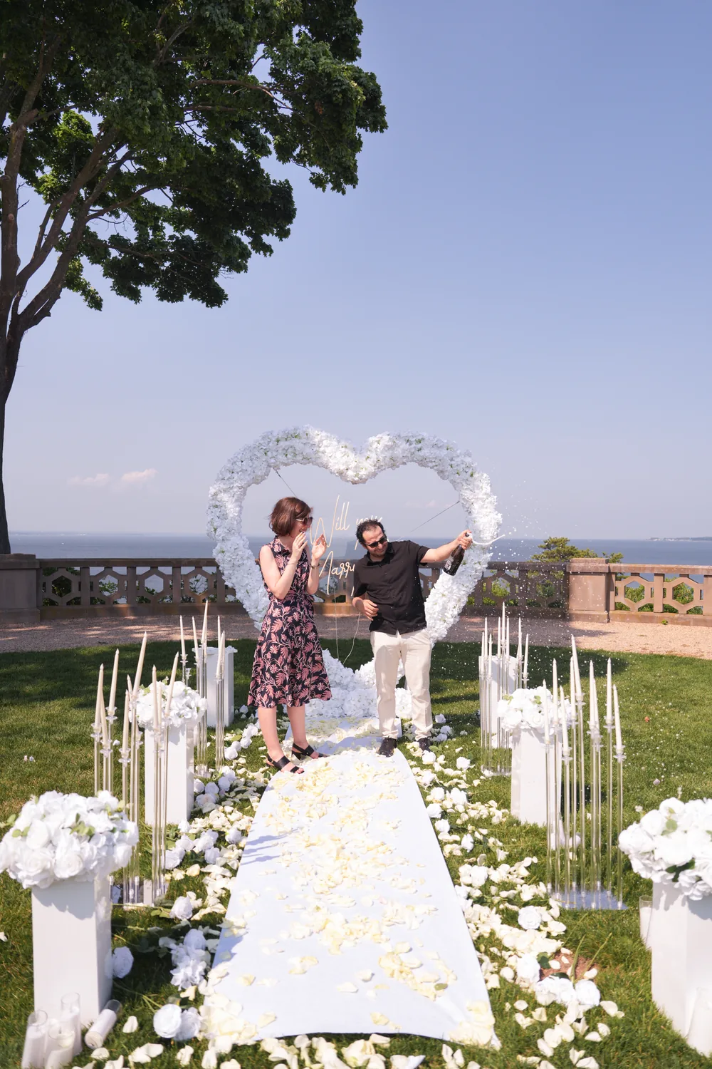 A couple under a white floral heart on a lawn by the water