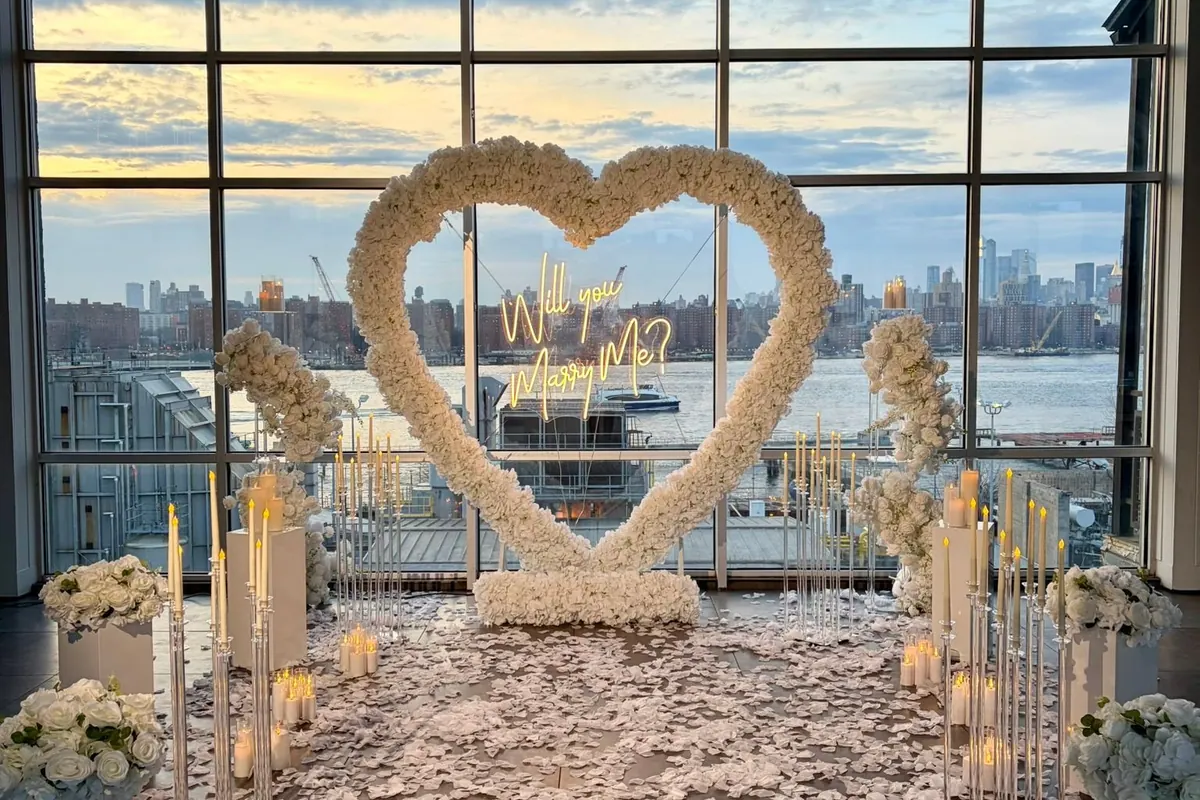 A white rose heart with a Will You Marry Me sign by tall windows overlooking the river, with candles and white petals