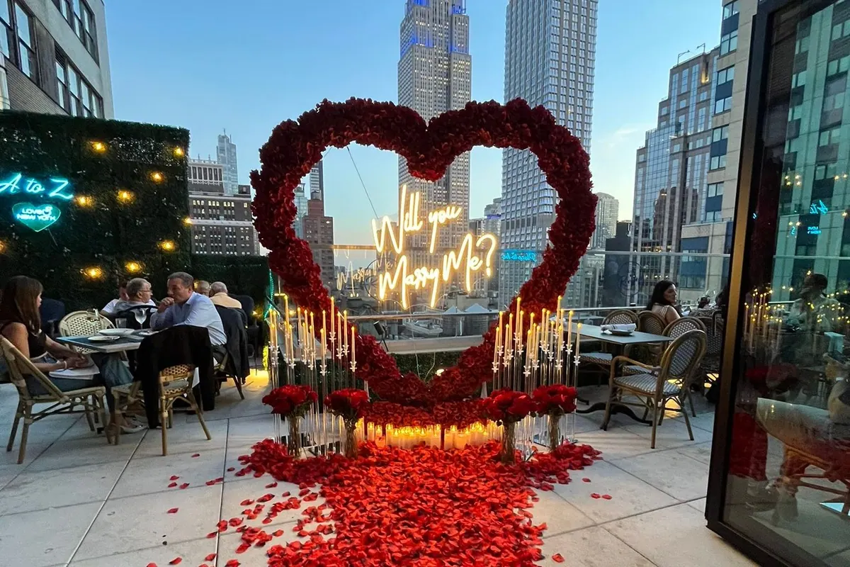 A red rose heart with a lit Will You Marry Me sign on a Manhattan rooftop terrace, the Empire State Building behind