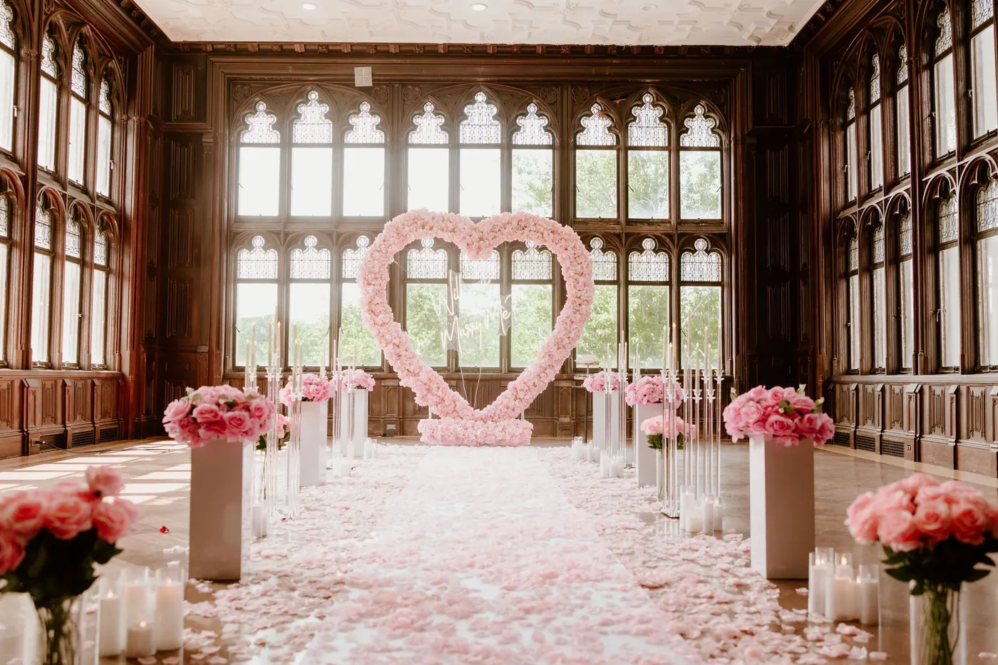 A pink rose heart at the end of a petal aisle lined with pink flowers and candles, in a hall of tall leaded windows