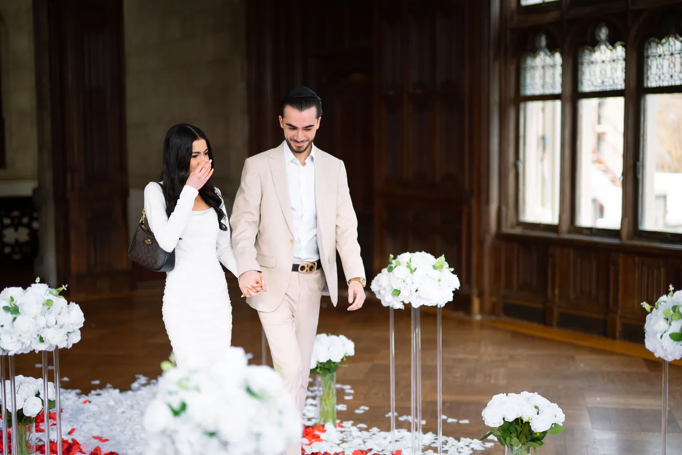 She walks into a wood-panelled hall of white flowers with a hand to her mouth as he walks beside her