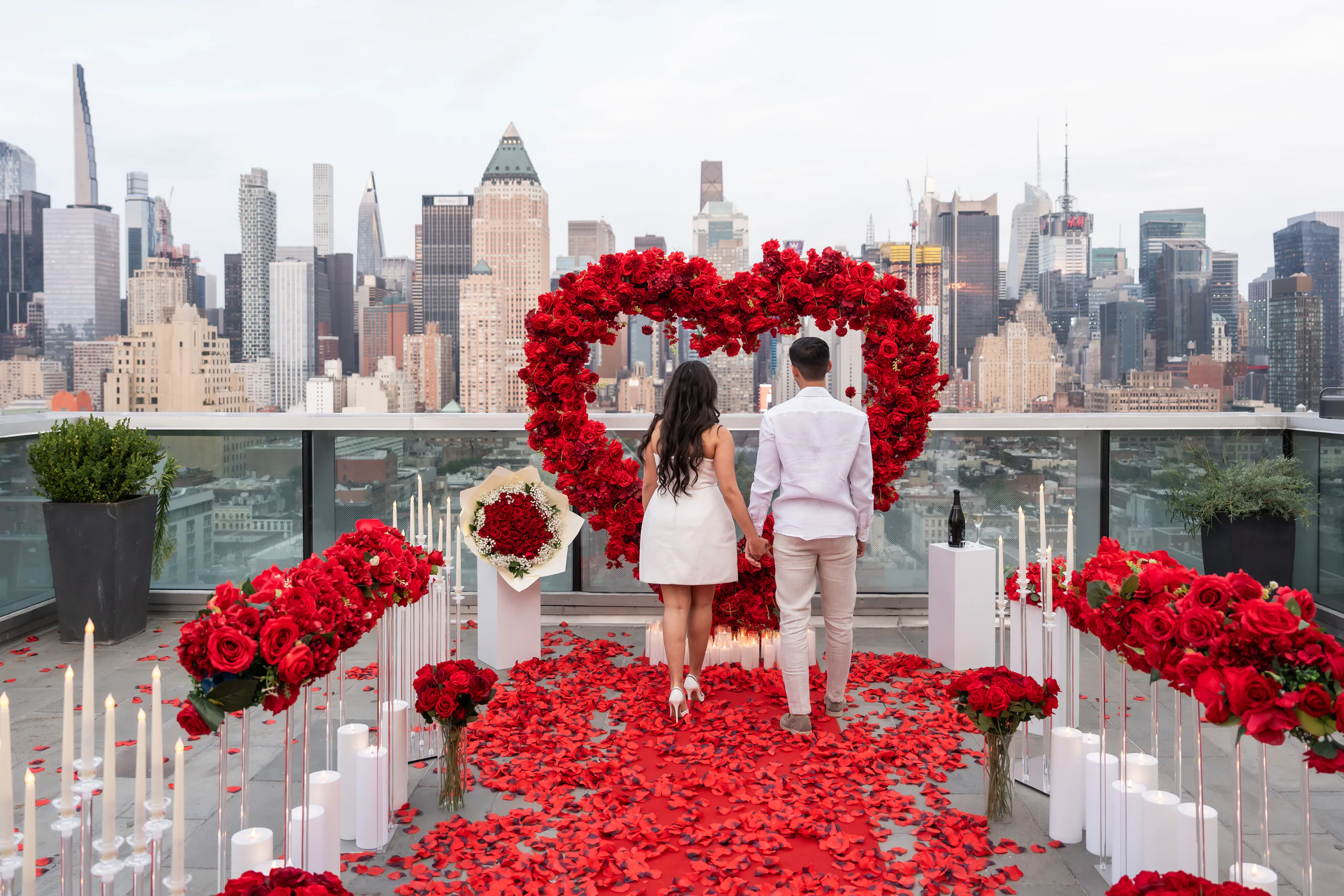 A couple seen from behind, standing inside a red rose heart on a Manhattan rooftop, looking out at the skyline