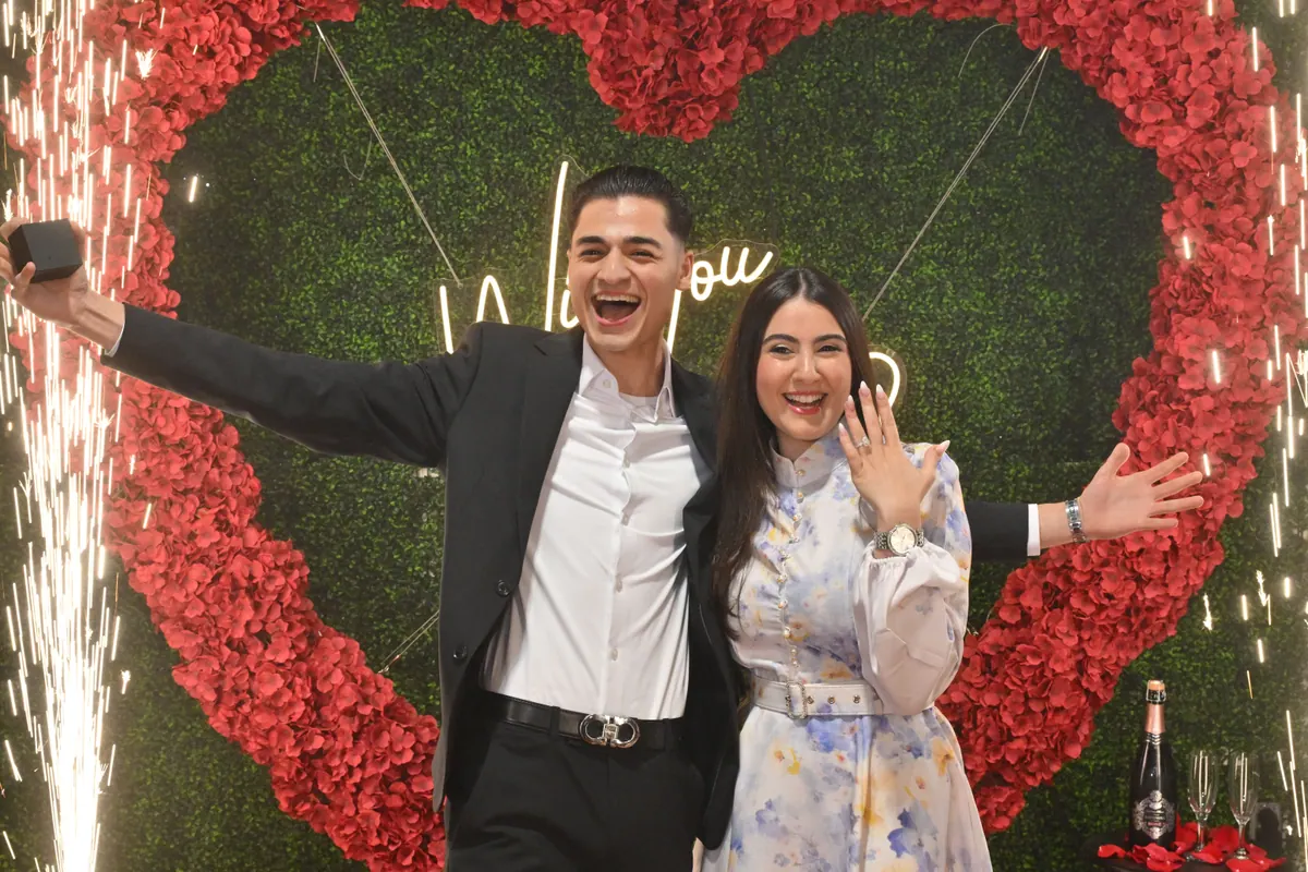 The couple cheer with arms raised in front of a red rose heart on a greenery wall