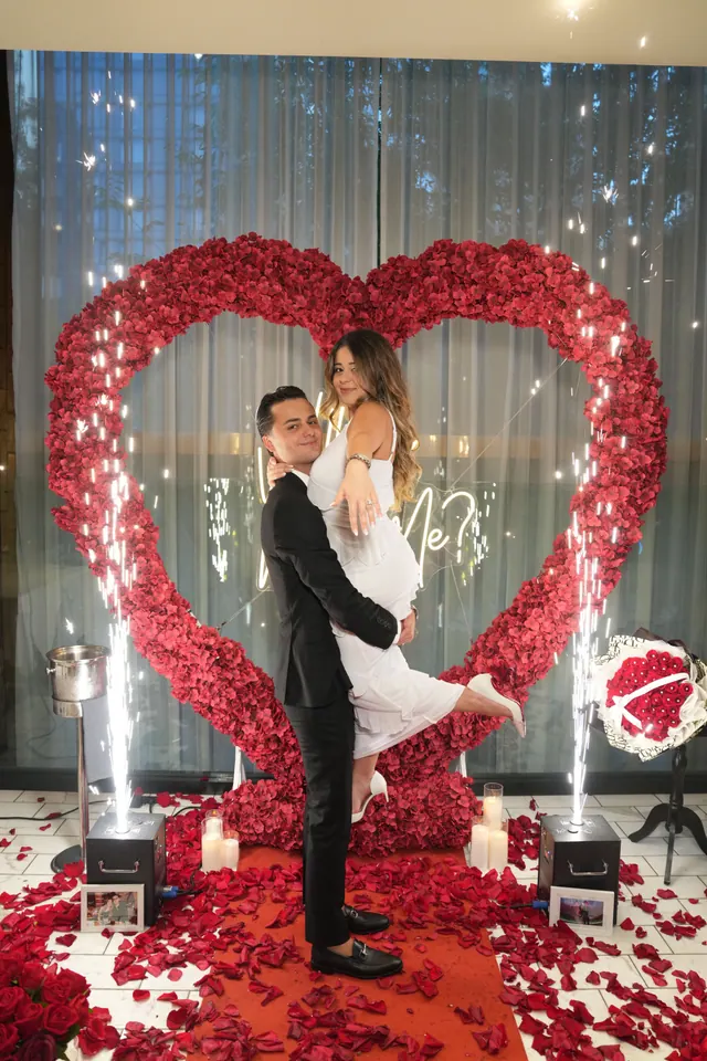 He lifts her inside a red rose heart as sparklers go off