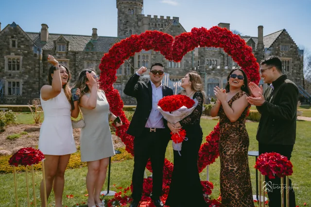 Friends cheering around the newly engaged couple in front of a red rose heart on a castle lawn