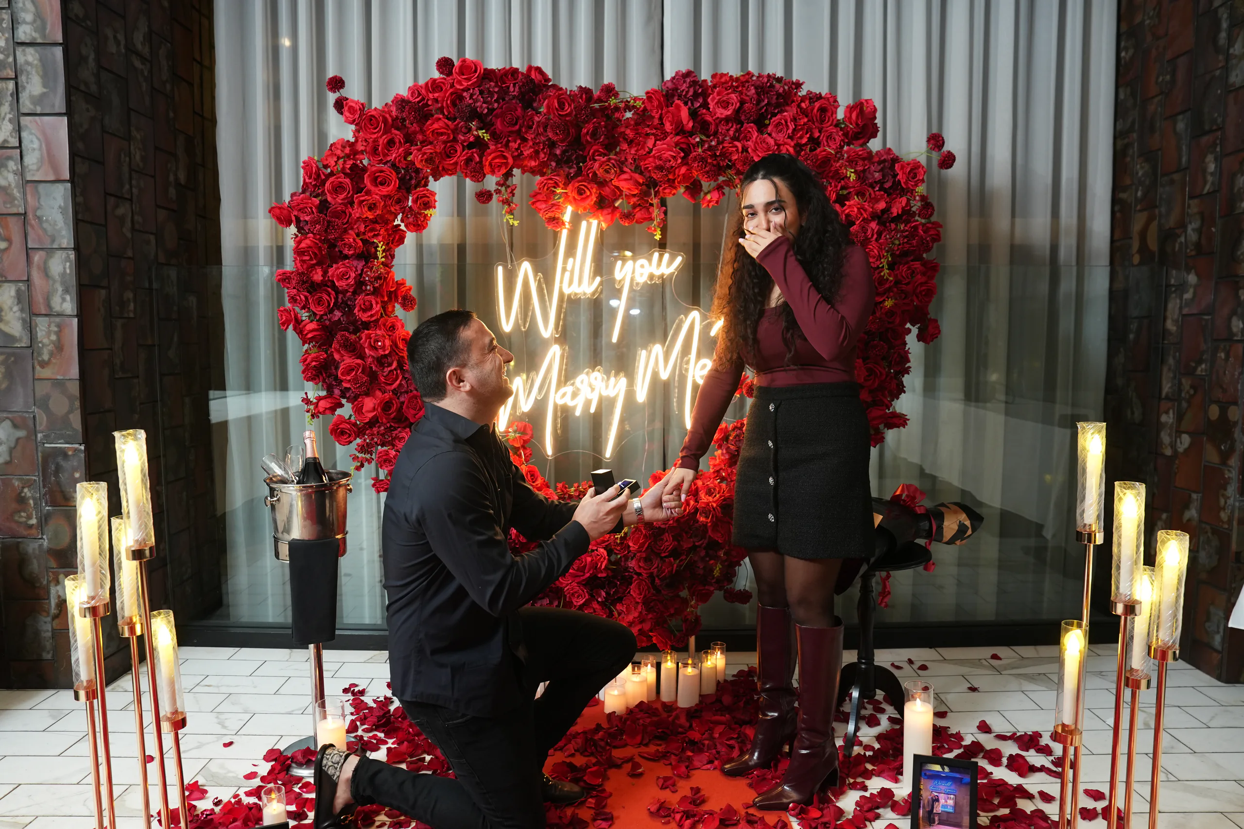 A man on one knee holding out a ring in front of a red rose heart with a glowing Will You Marry Me sign, as she reacts