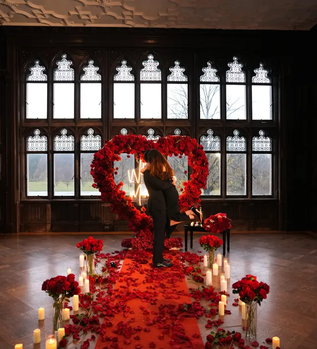 A man on one knee in a wood-panelled hall with leaded windows, a red rose heart and candlelit petal aisle behind him