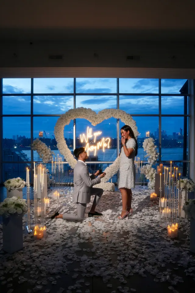 A man kneels before a white rose heart with a neon sign by windows at dusk as she covers her face