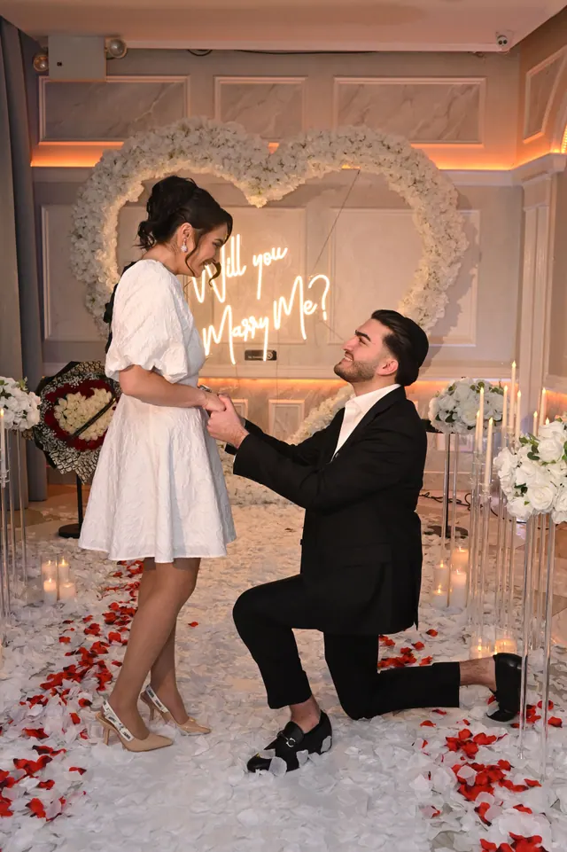 She laughs as he kneels on a floor of rose petals in a white room