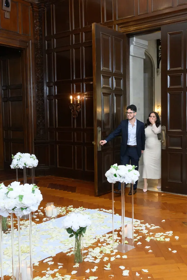 A couple stepping through tall wooden doors into a room of white flowers and petals