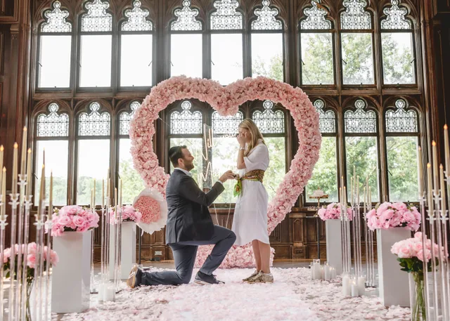 A man on one knee in front of a blush rose heart in a panelled hall as she stands facing him