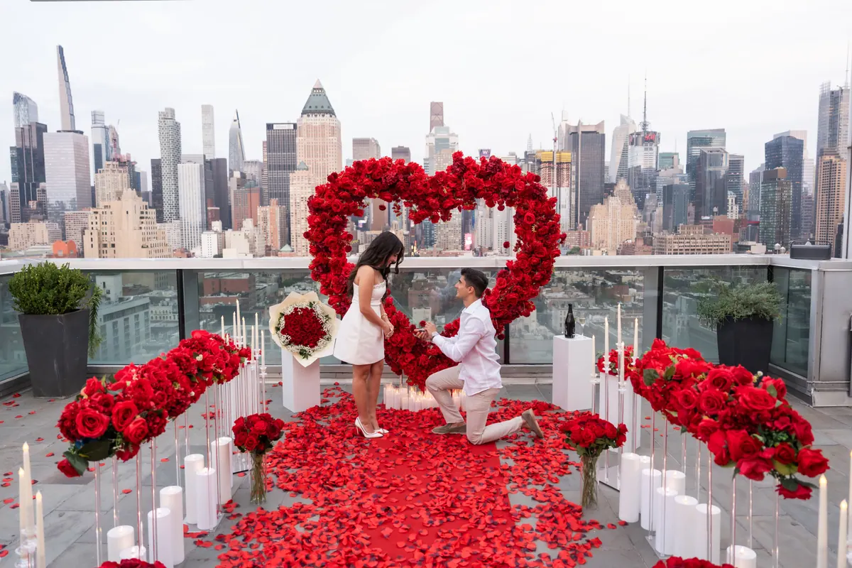 On a Manhattan rooftop, a man kneels inside an aisle of red roses as she turns to him, the skyline behind them