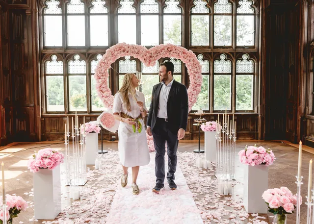 A newly engaged couple standing together in front of a blush rose heart, smiling at each other