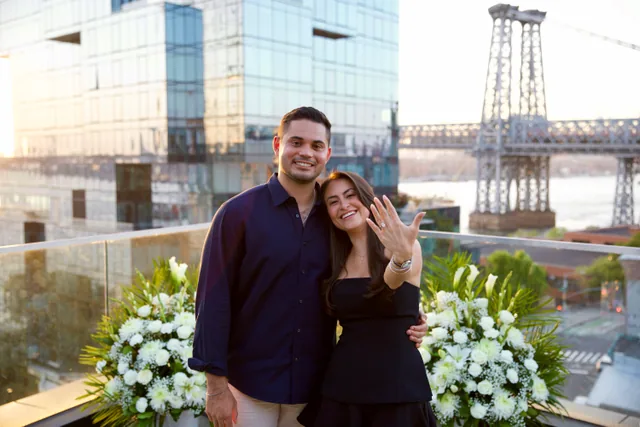 A woman holding up her hand to show the ring, leaning into her fiancé with the Williamsburg Bridge behind them