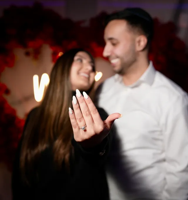A newly engaged woman holding up her ring, smiling at her fiancé in front of red roses
