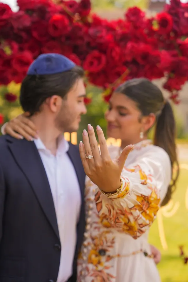 Smiling, she shows her ring to the camera beside him in front of red roses