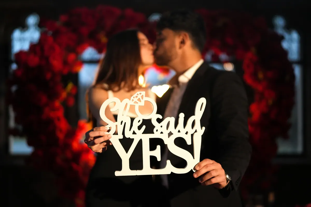 The couple kiss behind a She said YES sign in front of red roses