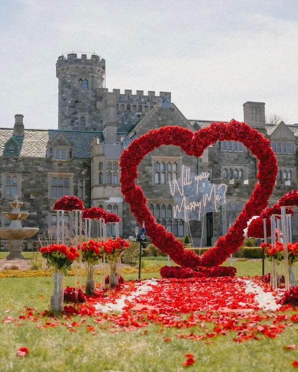 A red rose heart with a Will You Marry Me sign on the lawn in front of a stone castle