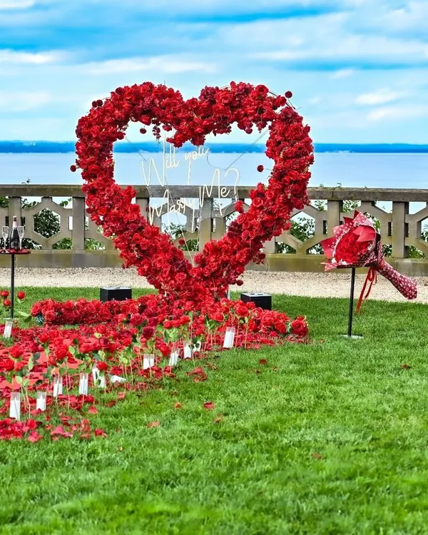 A red rose heart on a waterfront lawn beside a stone balustrade, with a winding path of petals