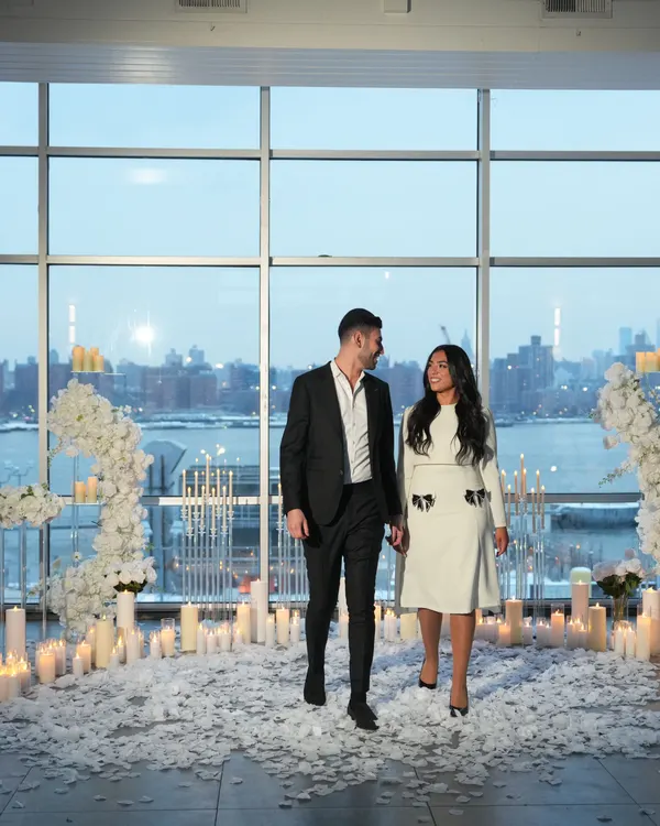 A couple walk in past lines of candles and white florals in a white room with river views