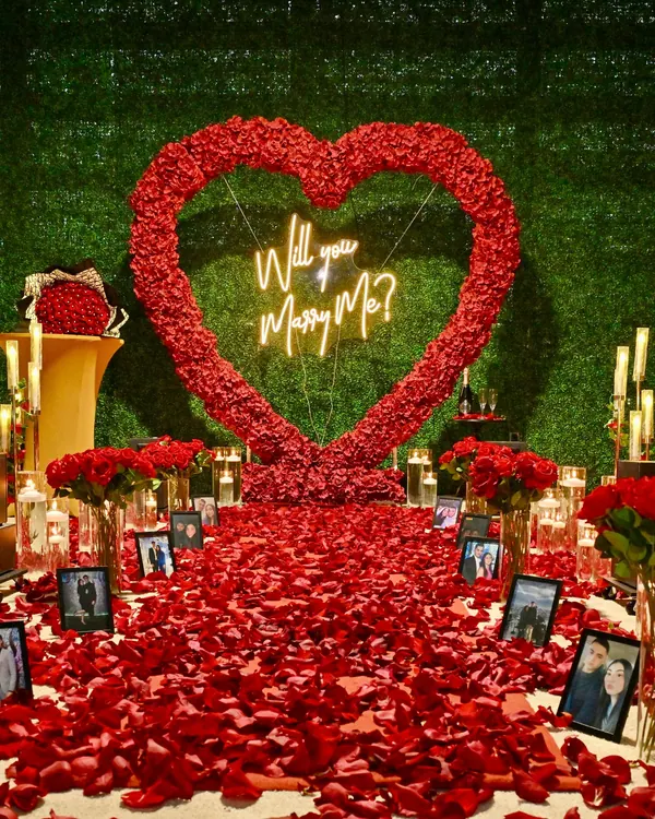 A red rose heart on a greenery wall with candles, roses and framed photographs of the couple