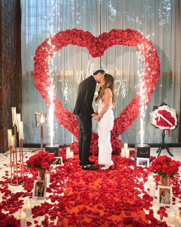 A couple kiss inside a red rose heart as sparklers go up either side, against a grey curtain