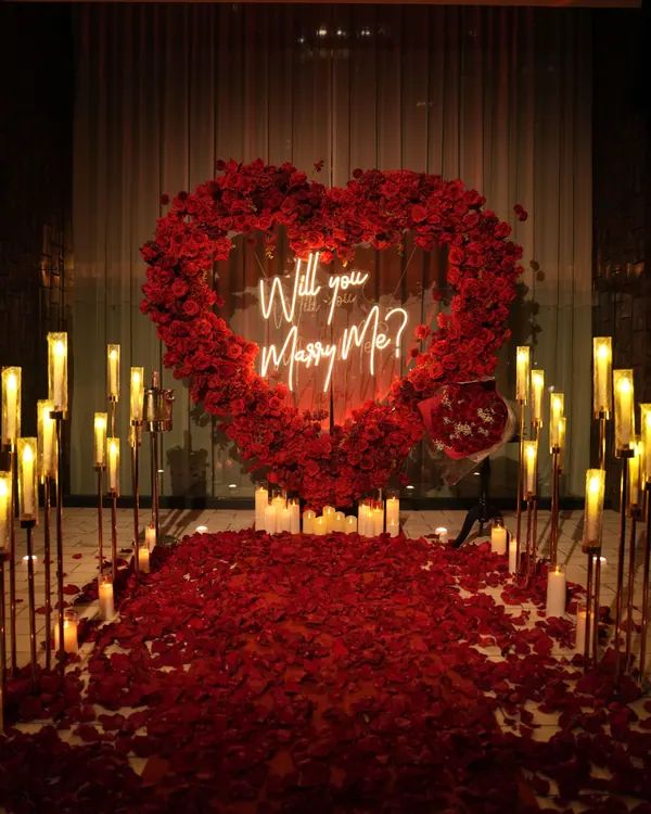 A red rose heart with a neon sign between tall gold candle stands over red petals