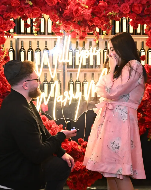 He kneels before a red rose wall with a neon sign behind a bar as she reacts