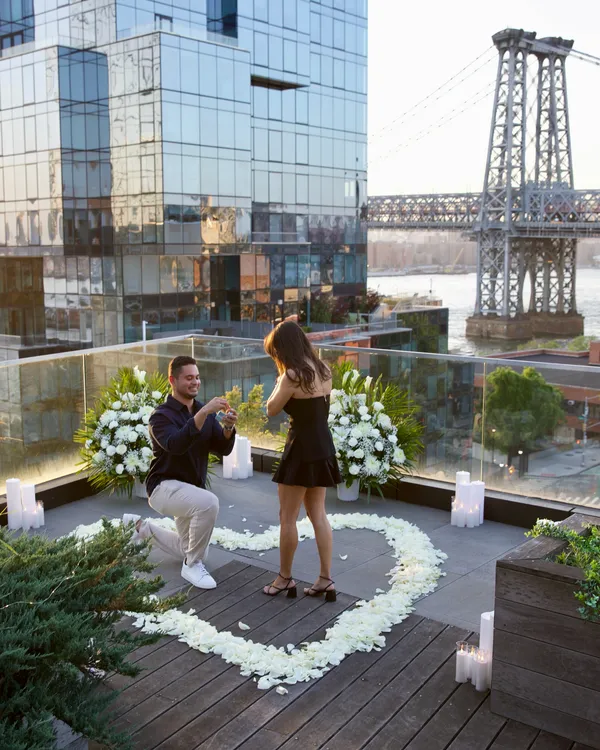 A proposal inside a heart of white petals on a planted roof deck, the Williamsburg Bridge behind