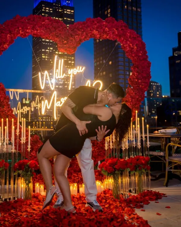 A couple kiss inside a red rose heart with a neon sign on a rooftop at night, between lit towers